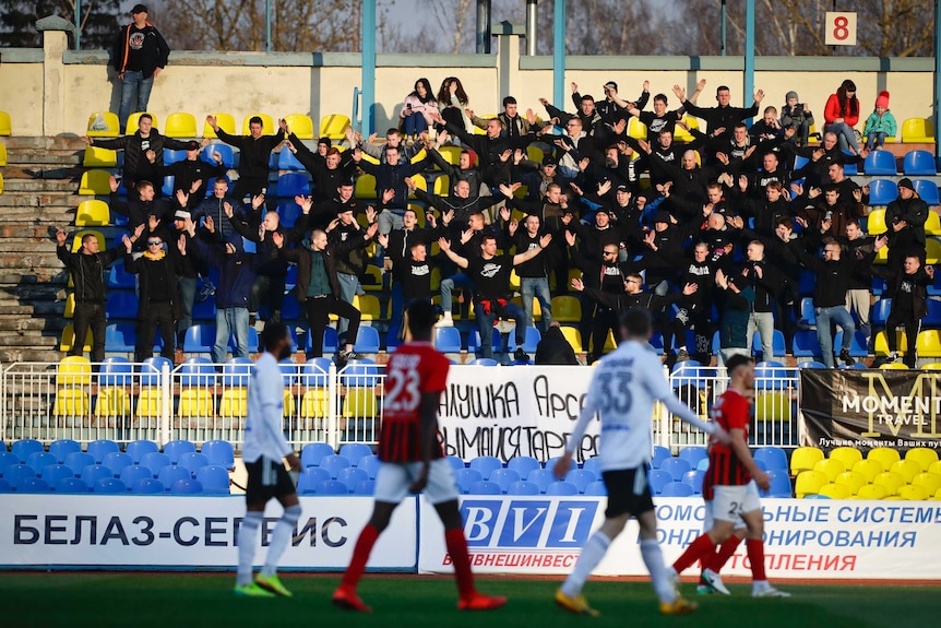 Fans stand with their arms outstretched in a grandstand with blue and yellow seats