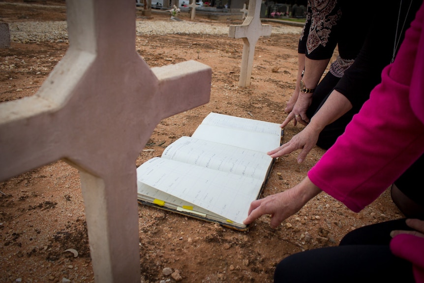 Old burial documents on the ground between graves with women's hands touching the pages.