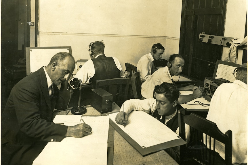 Black and white photo of six men at work on desks, writing on sheets, early radio equipment, old telephone.