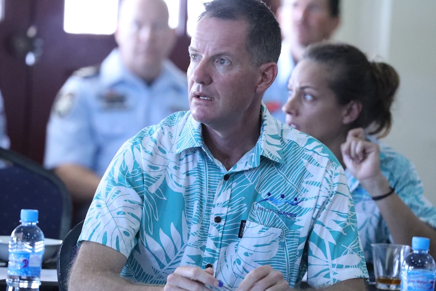 Man in green island print shirt stares to his right, appears to be in meeting room surrounded by people in same shirt. 