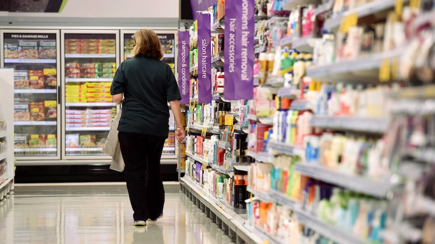 A woman walks down an aisle of a supermarket. Products on shelves either side of her.