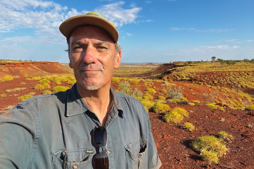 A selfie of a man wearing a cap with a desert landscape behind him. 