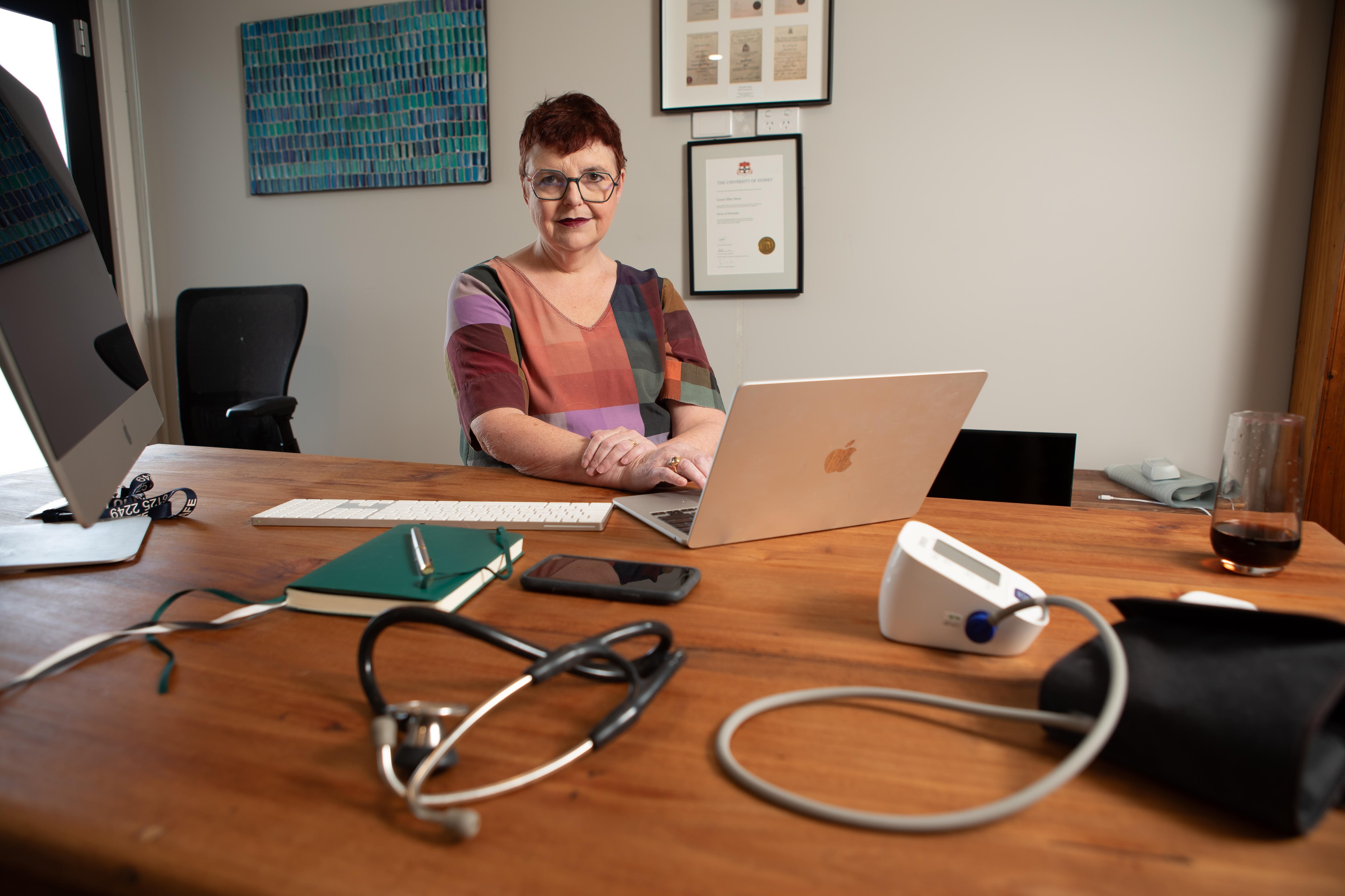 A wide angel shot of Louise who sits at a desk with a laptop and stethascope in front of her looking at the camera. 