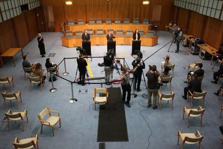 Aerial shot of three people behind podiums and camera crews, photographers and journalists spread out standing or on chairs.