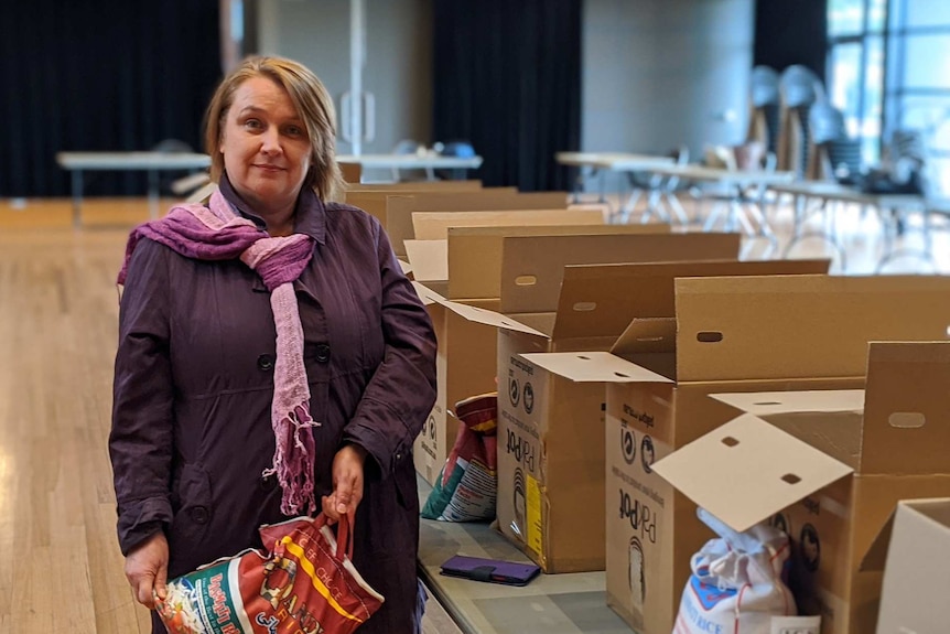 A woman in a purple jacket and scarf stands in a hall next to boxes on a table packed with food, she holds a large bag of rice.