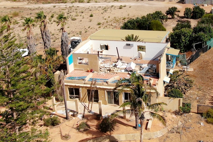 A two storey home with no roof and damage to the top floor which is covered with rubble. Vacant sandy land surrounds the home.