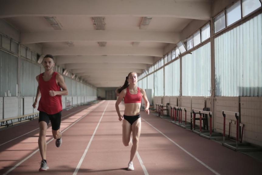 A man and woman run on a track indoors wearing red shirts and black pants.