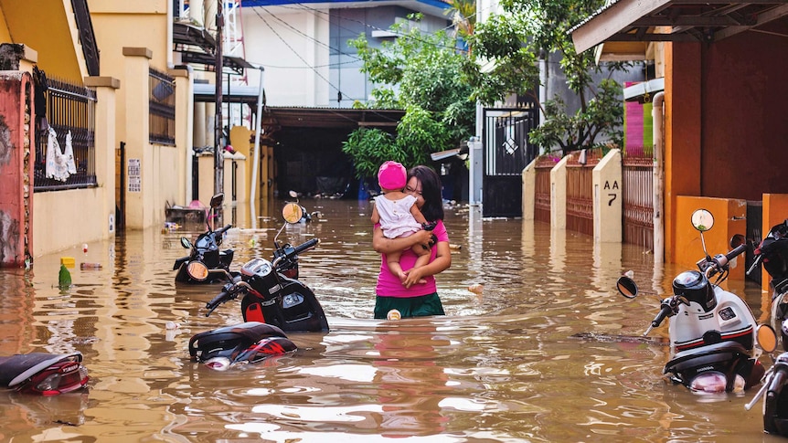 A woman wearing a pink shirt carries her daughter through floodwater in between two rows of houses.