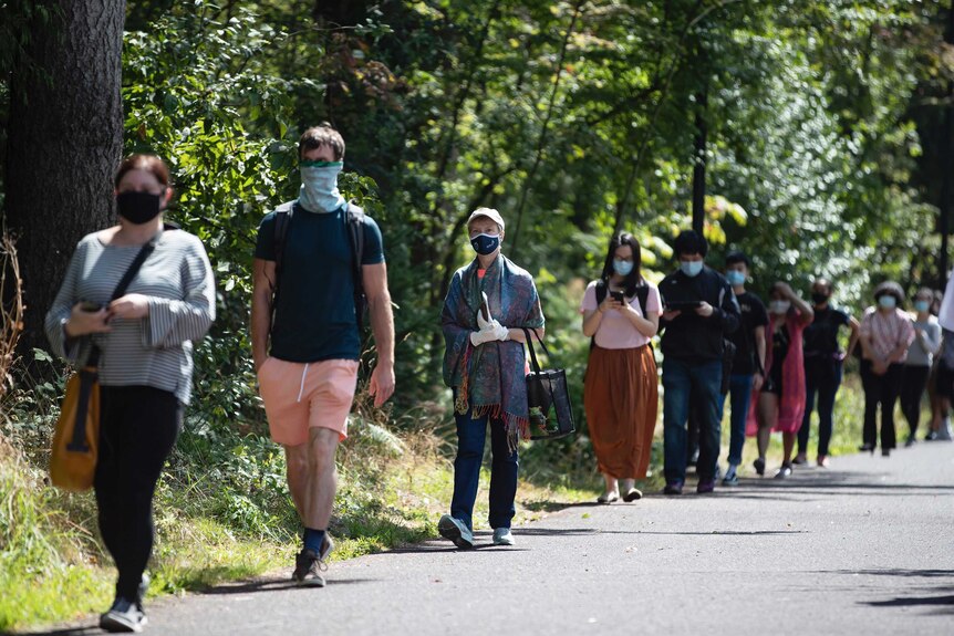 Men and women stand in line wearing masks in front of trees.