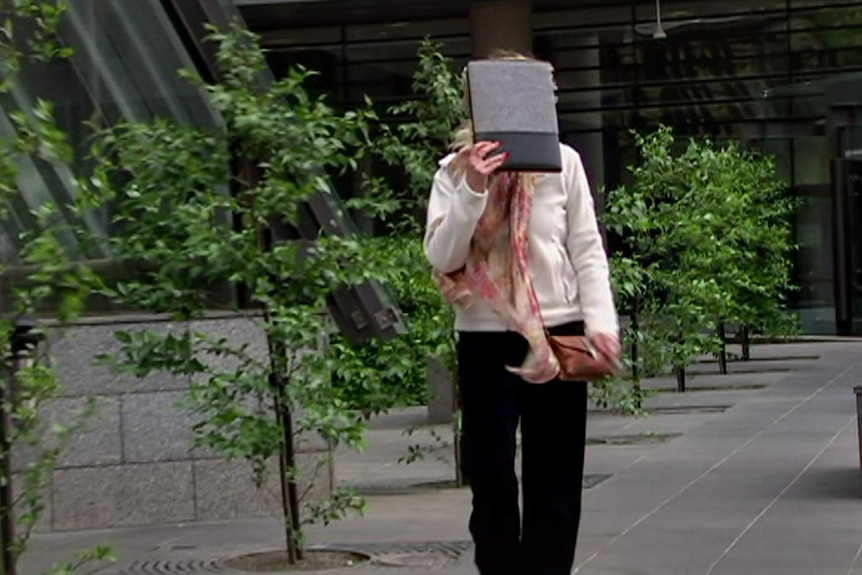A woman with white blouse, black pants and red painted fingernails, holds a book in front of her face as she walks.