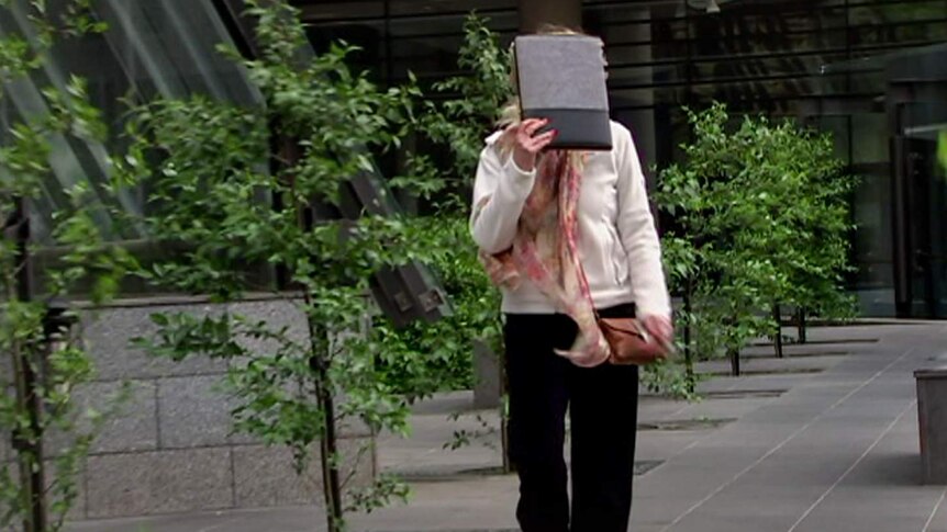 A woman with white blouse, black pants and red painted fingernails, holds a book in front of her face as she walks.