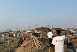 The aftermath of a cyclone with collapsed wooden houses and debris everywhere. Two men in white t-shirts are walking away.