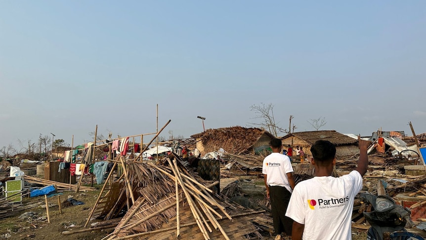The aftermath of a cyclone with collapsed wooden houses and debris everywhere. Two men in white t-shirts are walking away.