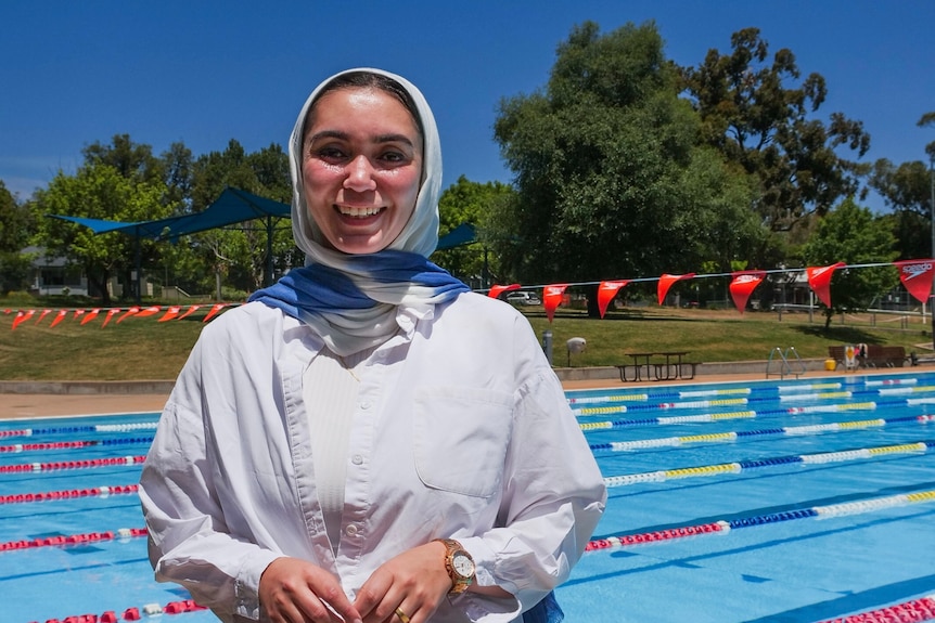 A woman in a white shirt and blue headscarf stands next to an outdoor 50 metre pool.