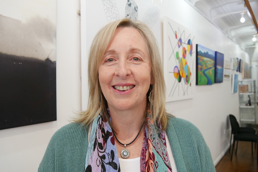 Woman with blonde hair and green jacket smiles at the camera, with artwork behind her 