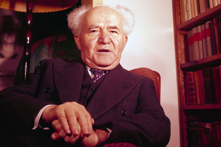 Israel's first Prime Minister David Ben-Gurion sitting, with palms on top of each other, with lamp and bookcase in background.