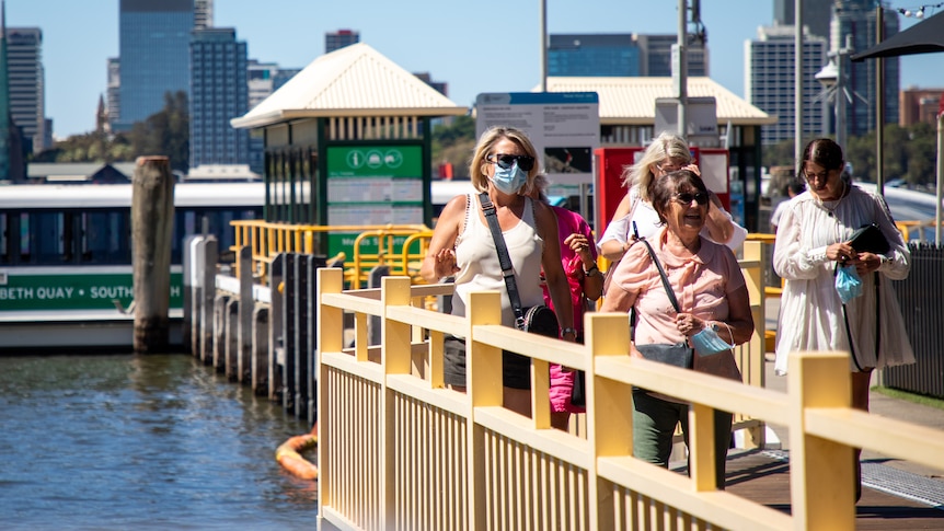 Passengers with face masks disembark a ferry in South Perth with the CBD and Swan River behind them.