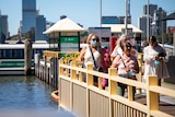 Passengers with face masks disembark a ferry in South Perth with the CBD and Swan River behind them.