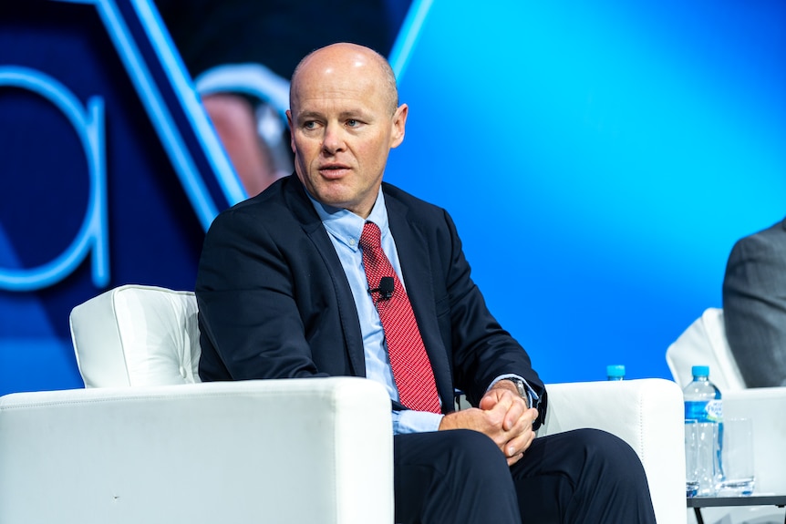 Man with shaved head wearing suit and tie, sitting in a chair and answering a question looking to his right