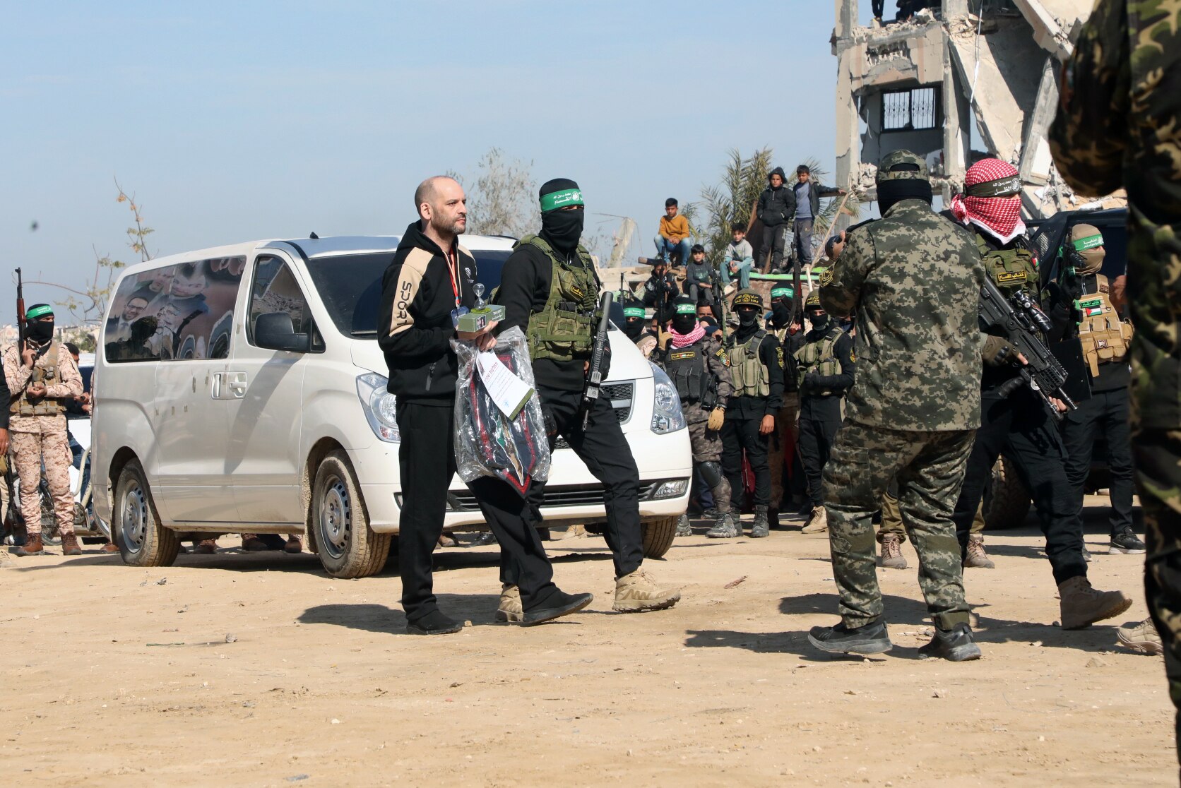 A balding white man walks in front of a white van escorted by a masked and armed militant 