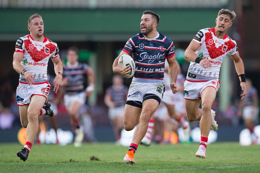 James Tedesco runs with the ball in one hand with two St George players in white shirts with a red v running after him.