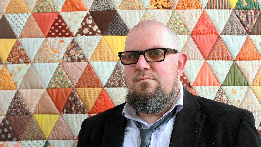 Man wearing glasses standing in front of patchwork quilt hanging on the wall.