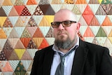 Man wearing glasses standing in front of patchwork quilt hanging on the wall.