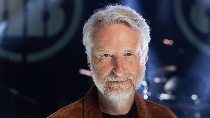 Billy Bragg, a man with white hair and beard, smiles directly into camera with a dark stage behind him