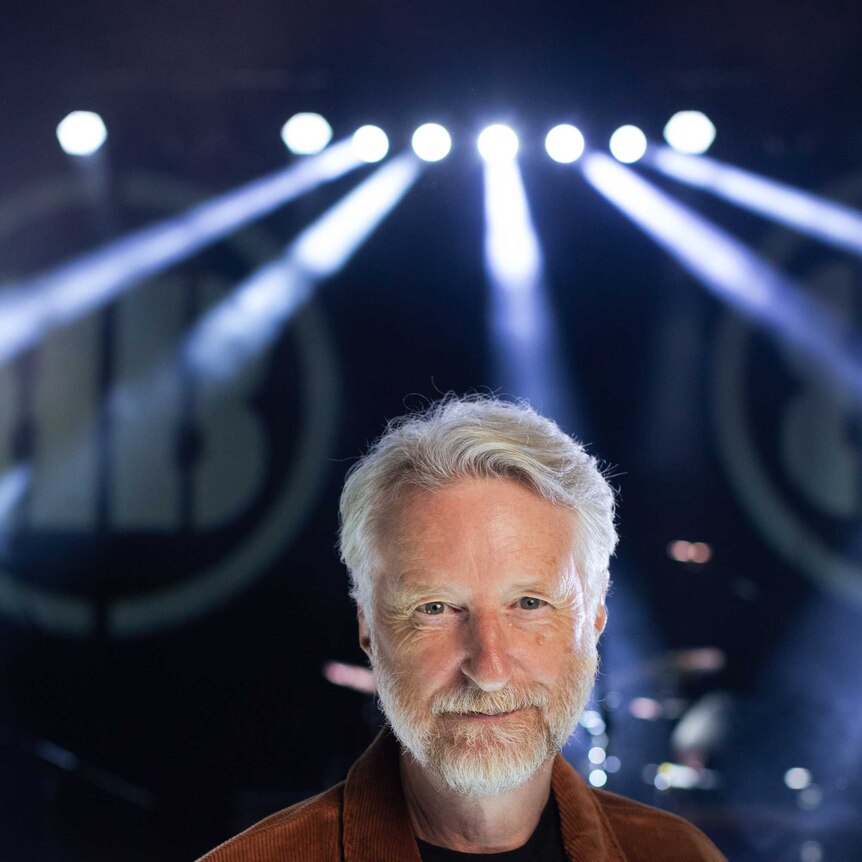 Billy Bragg, a man with white hair and beard, smiles directly into camera with a dark stage behind him