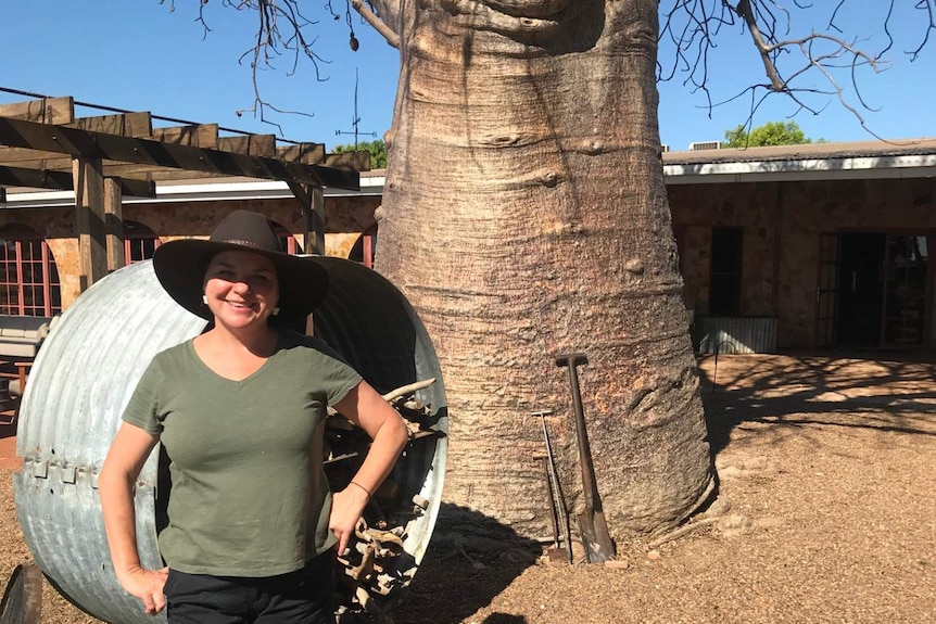 woman wearing hat smiling standing in front of large boab in homestead gravel courtyard. Blue skies