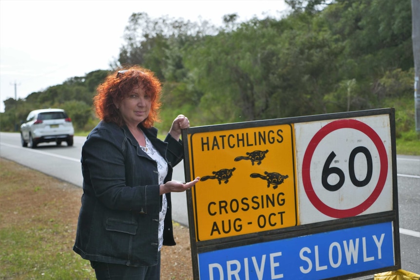 A woman stands next to a roadside sign holding a freshwater turtle hatchling.