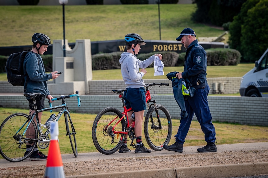 Two boys on bikes, one holding a piece of paper up as a policeman approaches.