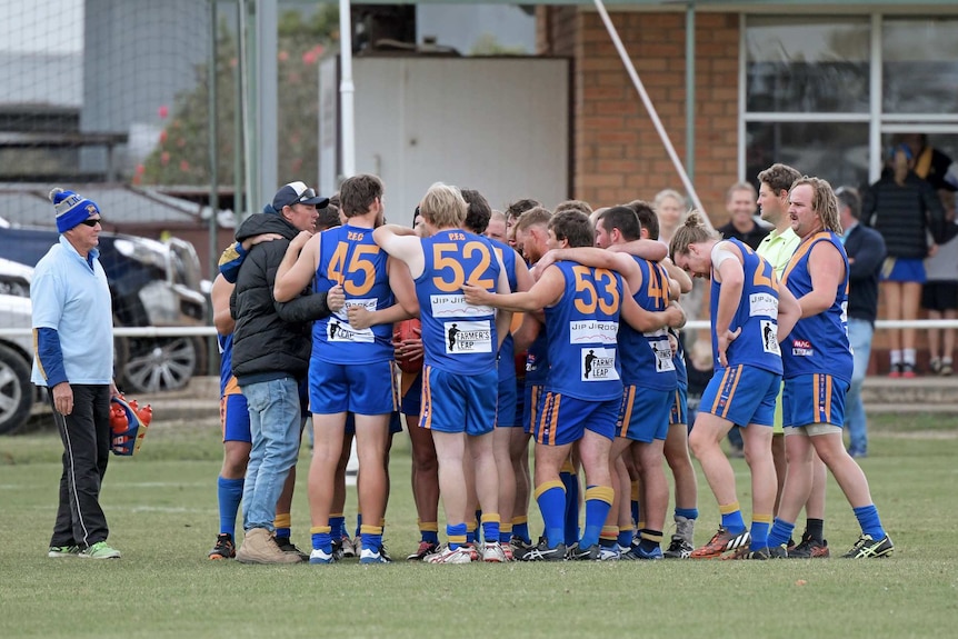 A group of men footballers in blue and gold jumpers huddle with their arms around one another