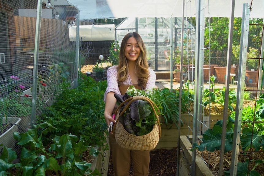 A woman in an overflowing veggie garden holds out a basket of produce.