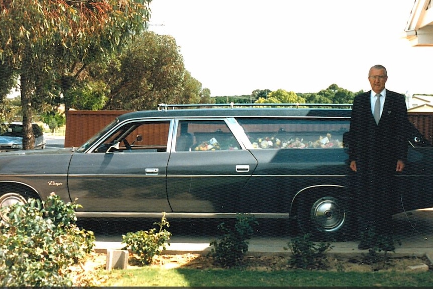 A man in a suit stands at the side of a hearse with a coffin in it.