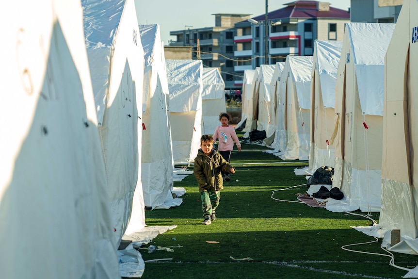 A boy and a girl run on grass between white tents.