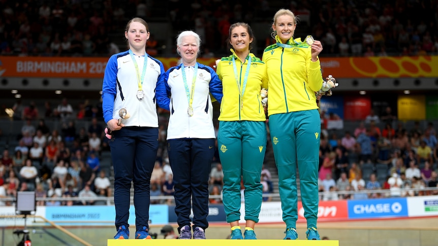 Four women, two wearing white and blue and two wearing yellow and green, stand on a podium