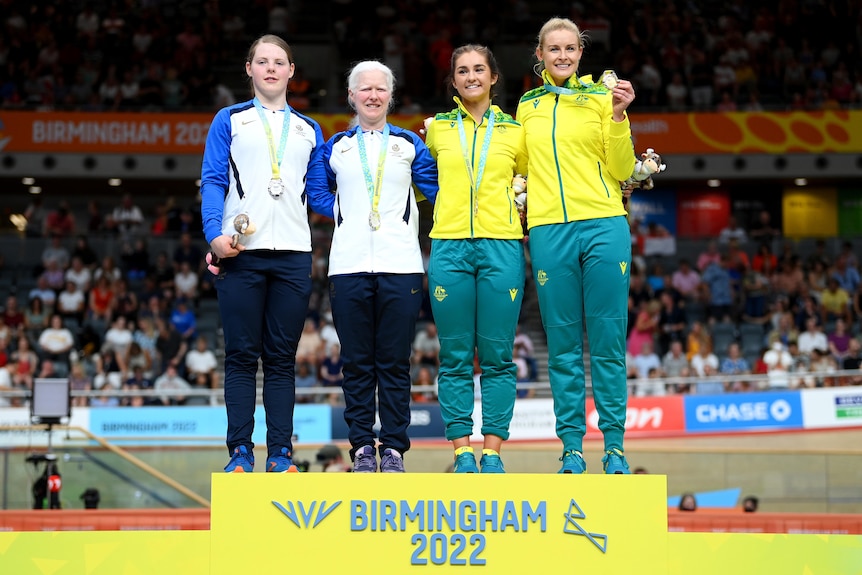 Four women, two wearing white and blue and two wearing yellow and green, stand on a podium