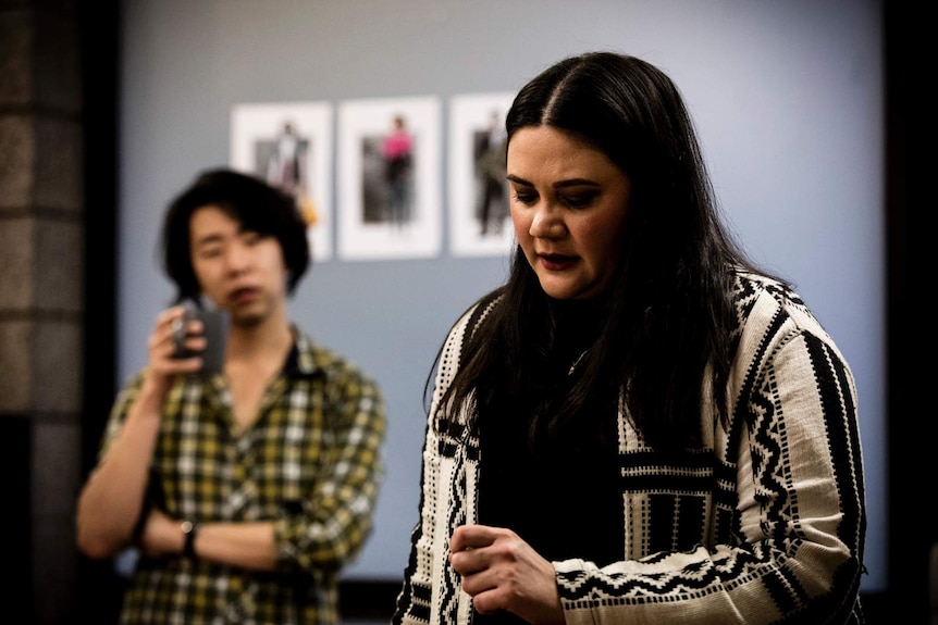 Shari Sebbens looking downward. A person holding a mug is out of focus behind her.