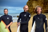 Three surfers at Bells Beach wearing wetsuits and carrying surfboards smile at the camera.