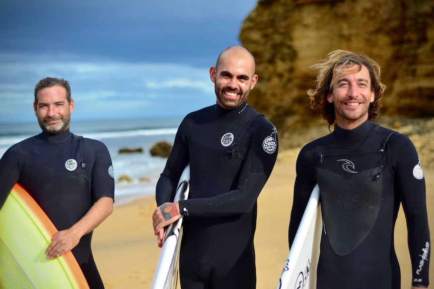Three surfers at Bells Beach wearing wetsuits and carrying surfboards smile at the camera.