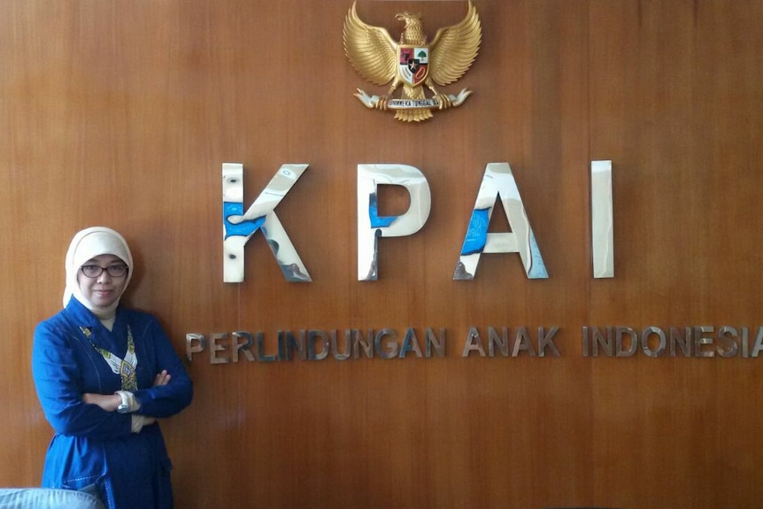 A woman leaning against a wooden wall with "KPAI" in silver lettering under a golden eagle statue