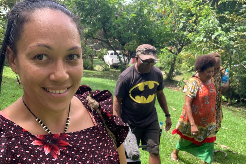 A woman in the foreground smiles while walking with a group on a grassy, tree-lined path.