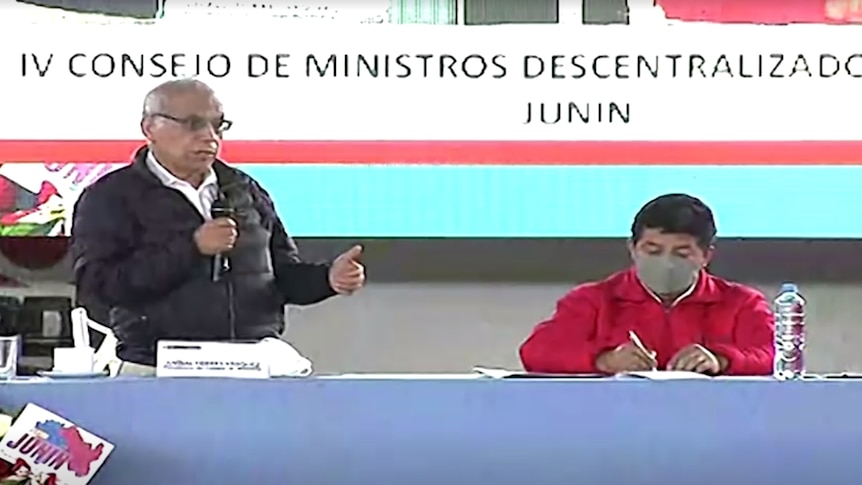 A man holding a microphone gestures while speaking behind a table with a person seated next to him taking notes.