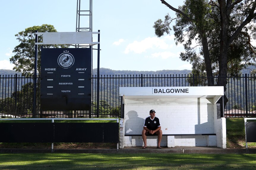 Steve Buckley sits inside a white dugout next to a scoreboard, looking off camera.