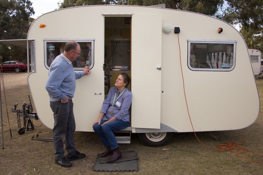 A woman sits on the steps to a small vintage caravan and looks up at her husband, who is leaning on the side of the van.