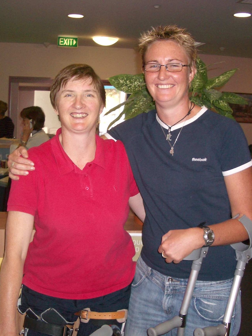 Woman in red shirt in arms with woman with dark blue t-shirt. Wrist crutches hanging off her arm. Both women smiling.