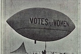 Black and white photograph of an airship flying over trees in London bearing the slogan Votes for Women.