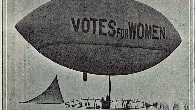 Black and white photograph of an airship flying over trees in London bearing the slogan Votes for Women.