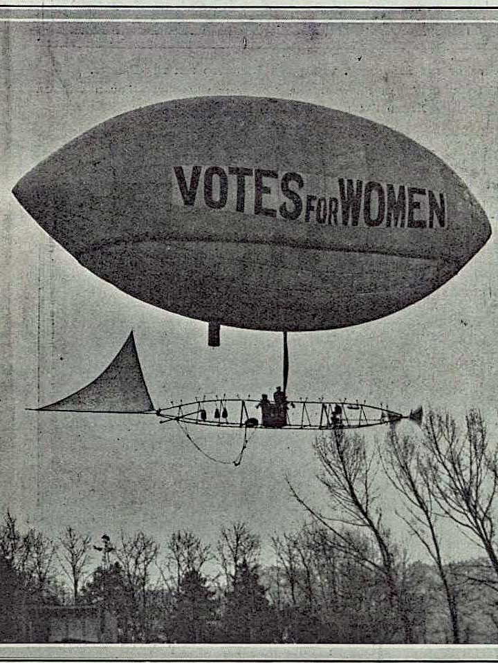 Black and white photograph of an airship flying over trees in London bearing the slogan Votes for Women.